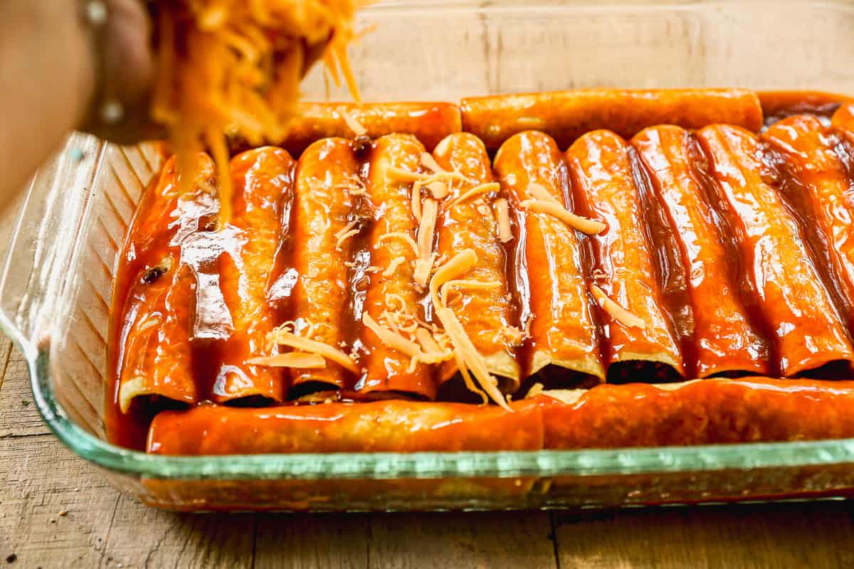 A hand sprinkles shredded cheese over rolled ground beef enchiladas covered in red sauce in a glass baking dish.