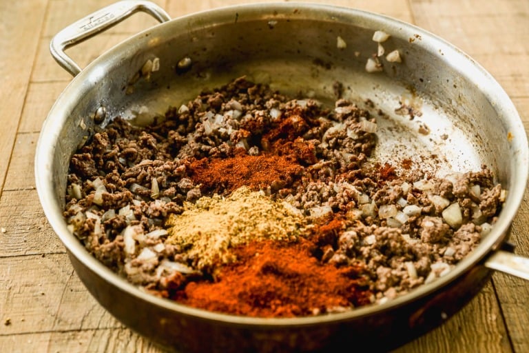 Ground beef and chopped onions cooking in a pan with piles of chili powder and other spices on top.