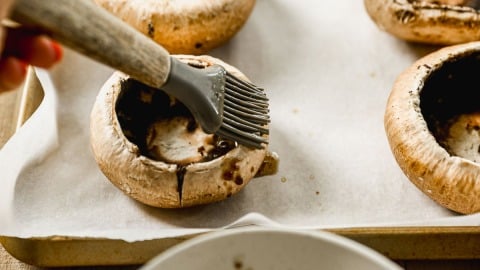A hand brushes marinade onto large portobello mushroom caps on a parchment-lined baking sheet.
