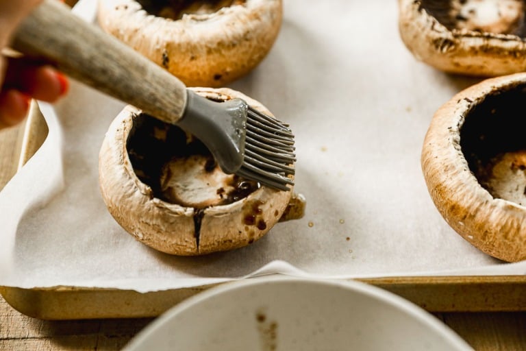 A hand brushes marinade onto large portobello mushroom caps on a parchment-lined baking sheet.