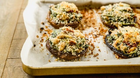 Four stuffed portobello mushrooms with a topping of breadcrumbs and herbs on a parchment-lined baking tray.