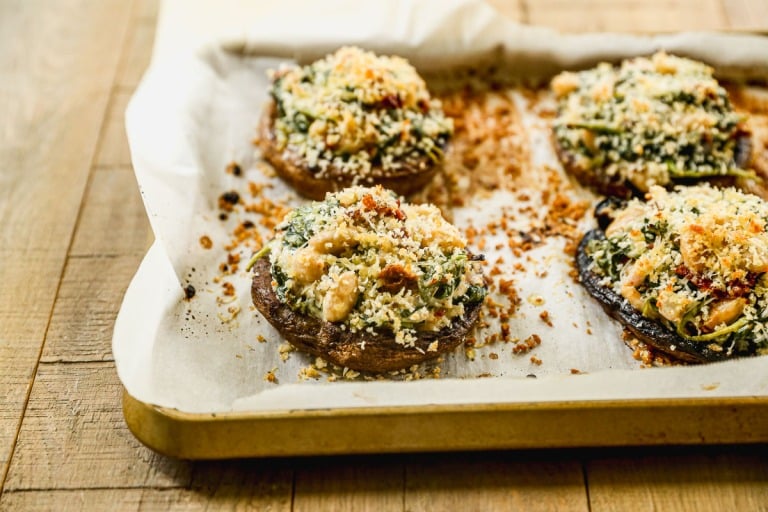 Four stuffed portobello mushrooms with a topping of breadcrumbs and herbs on a parchment-lined baking tray.