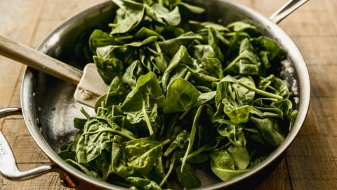 Fresh spinach leaves being sautéed in a stainless steel pan with a white spatula on a wooden surface.