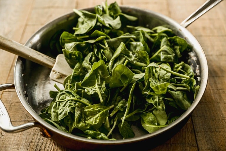 Fresh spinach leaves being sautéed in a stainless steel pan with a white spatula on a wooden surface.