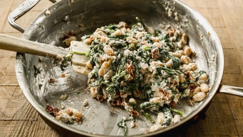 A skillet with creamy spinach, white beans, and sun-dried tomatoes being stirred with a spatula on a wooden surface.