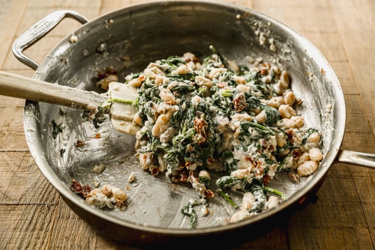 A skillet with creamy spinach, white beans, and sun-dried tomatoes being stirred with a spatula on a wooden surface.