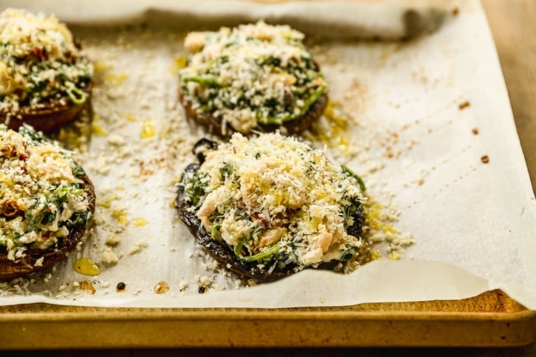 Stuffed portobello mushrooms topped with breadcrumbs and greens on a parchment-lined baking sheet.