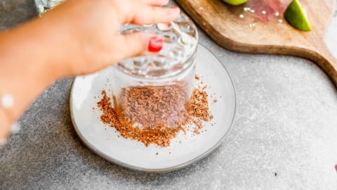 A hand twists a glass in Tajin on a plate, with lime wedges on a cutting board nearby.