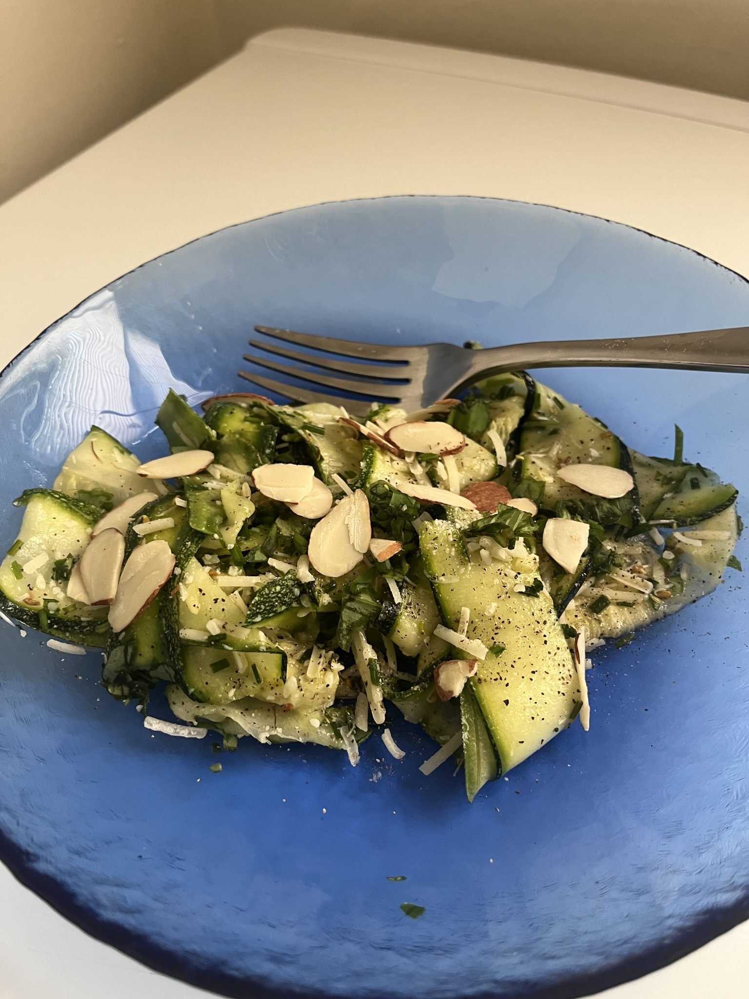 Zucchini ribbon salad with sliced almonds and grated cheese in a blue bowl, served with a fork.