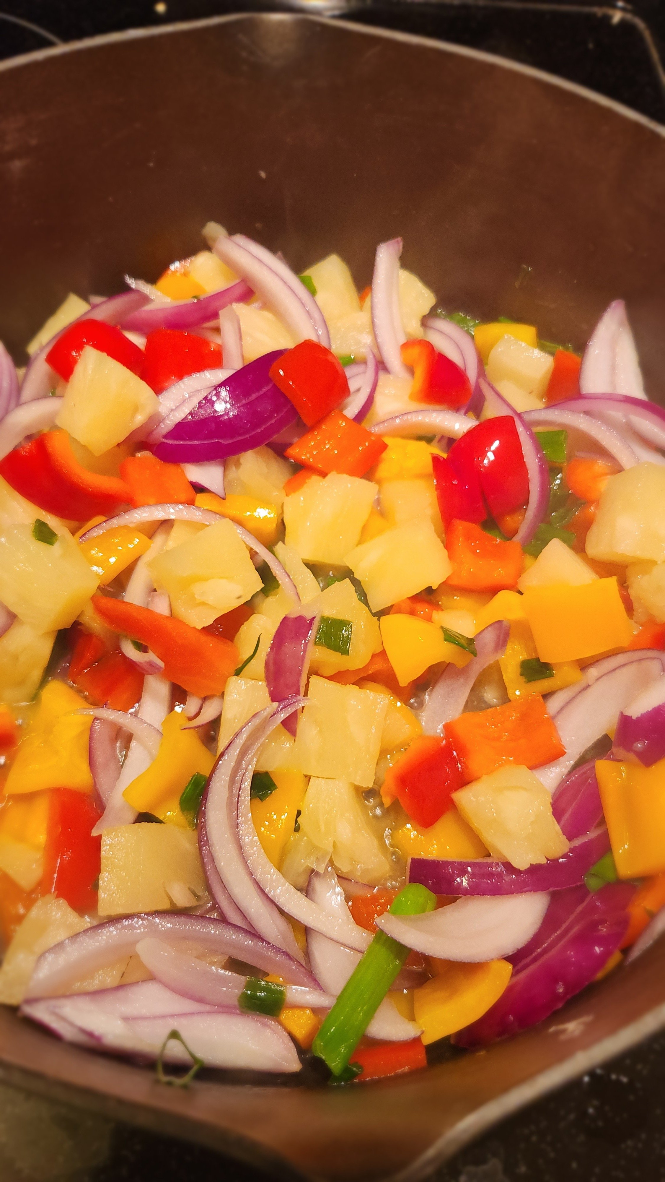 Colorful stir-fry with red onions, bell peppers, pineapple, and green onions cooking in a pan.