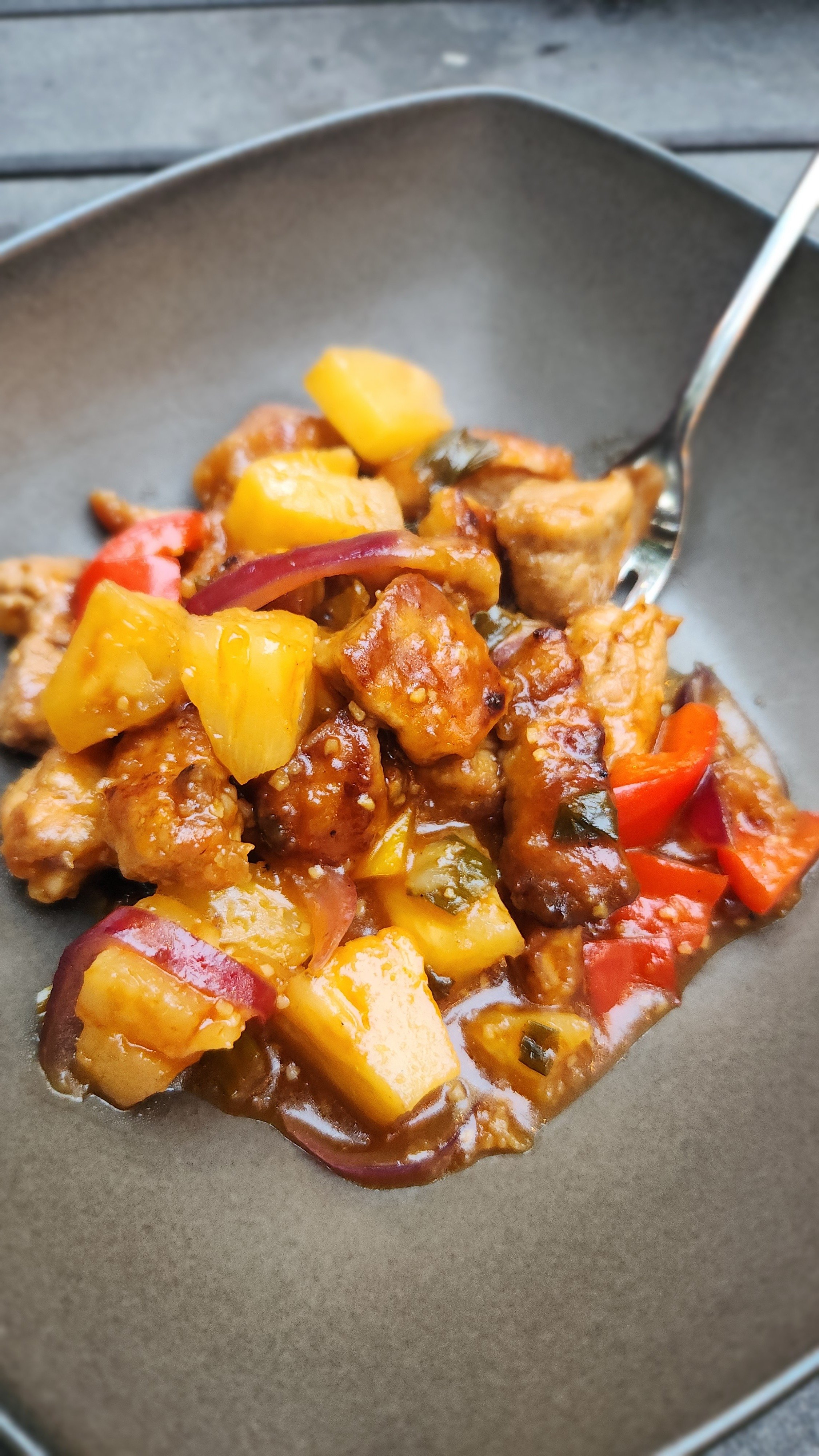 A plate of sweet and sour pork with pineapple, red bell peppers, and onions, served with a silver spoon.