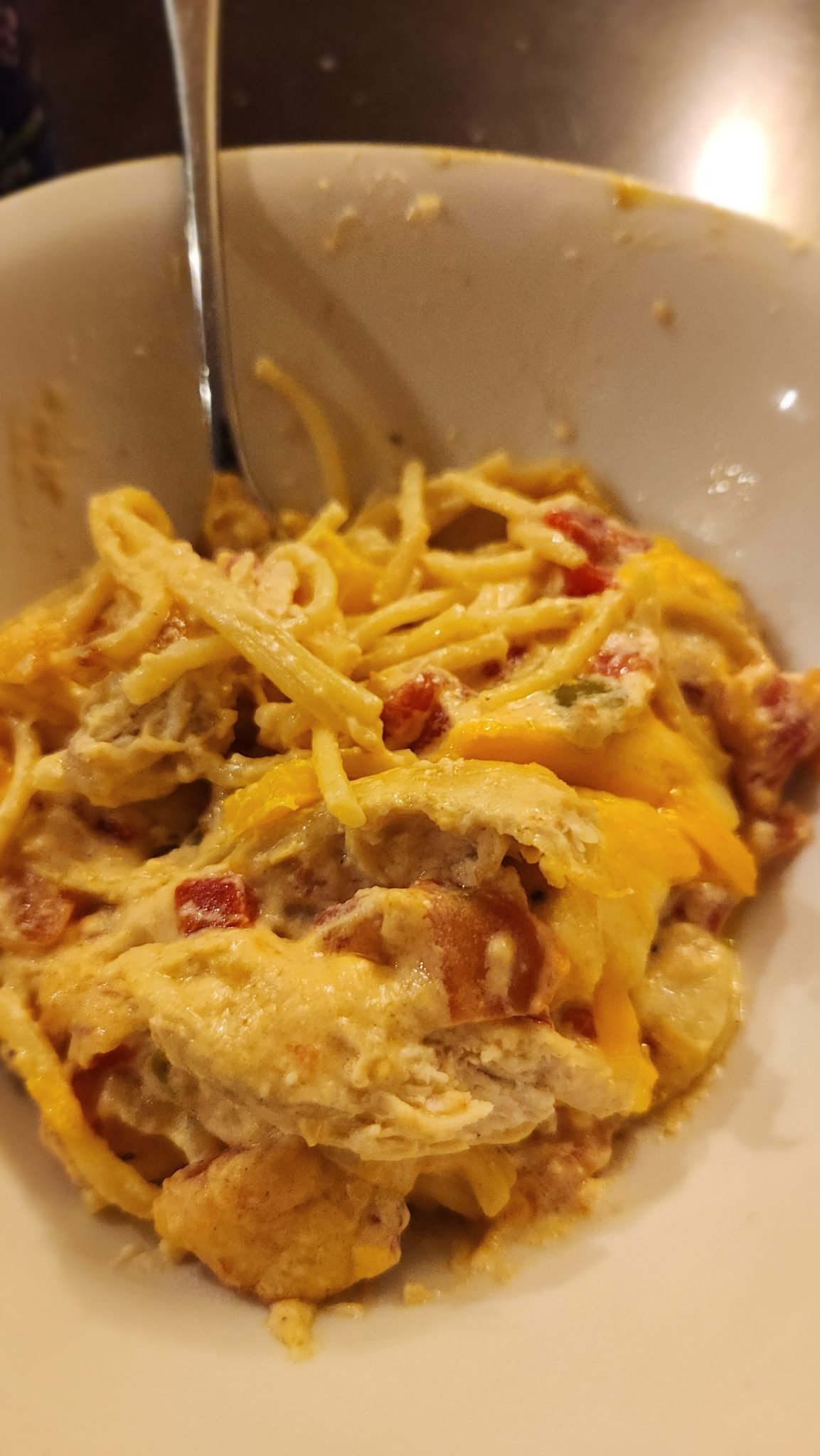 A bowl of cheesy spaghetti mixed with chunks of chicken and diced tomatoes, with a fork resting inside.