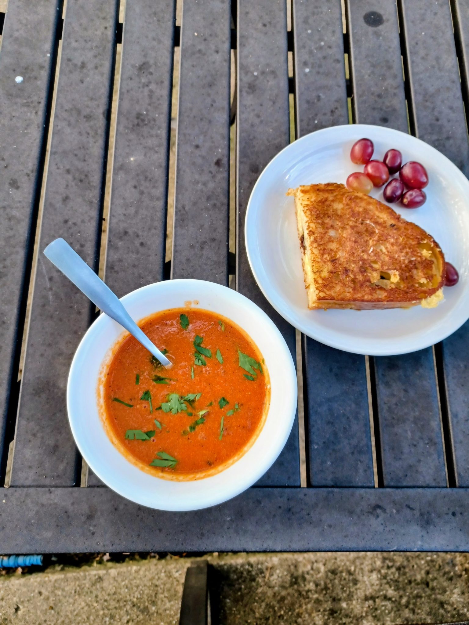 Bowl of tomato soup with herbs, grilled cheese sandwich, and grapes on a wooden table.