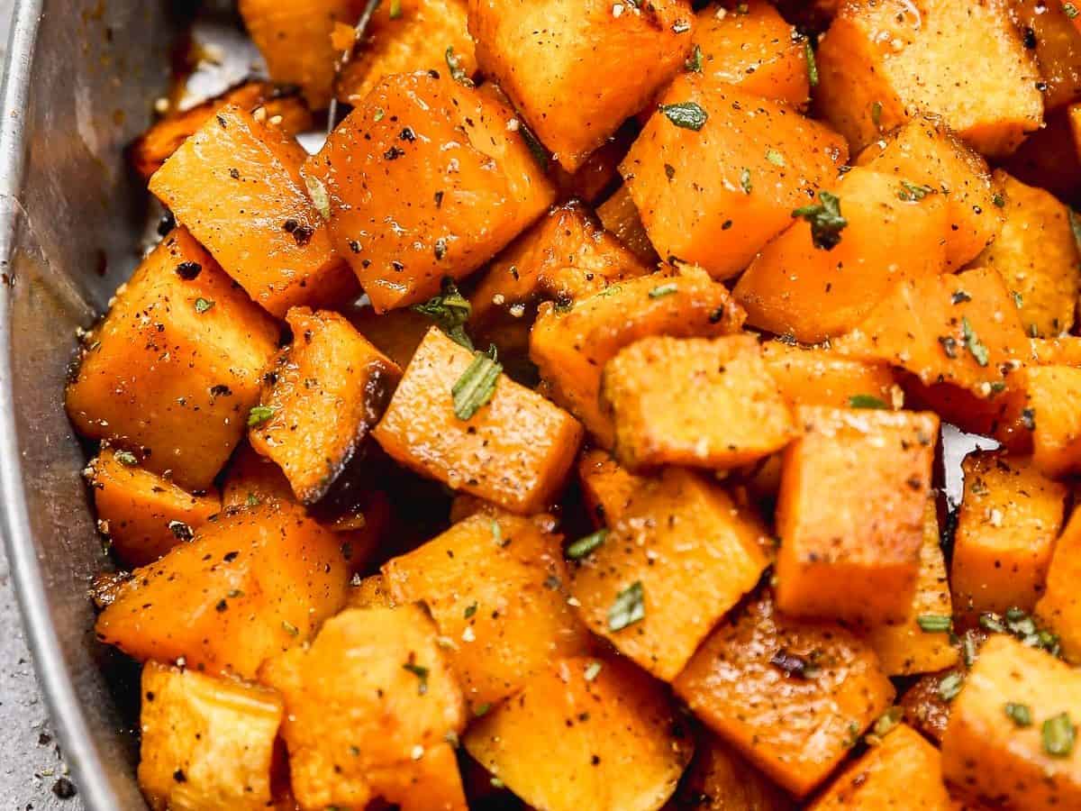 A close-up of roasted butternut squash cubes seasoned with herbs and pepper in an oval dish with a spoon.