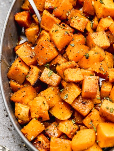 A close-up of roasted butternut squash cubes seasoned with herbs and pepper in an oval dish with a spoon.
