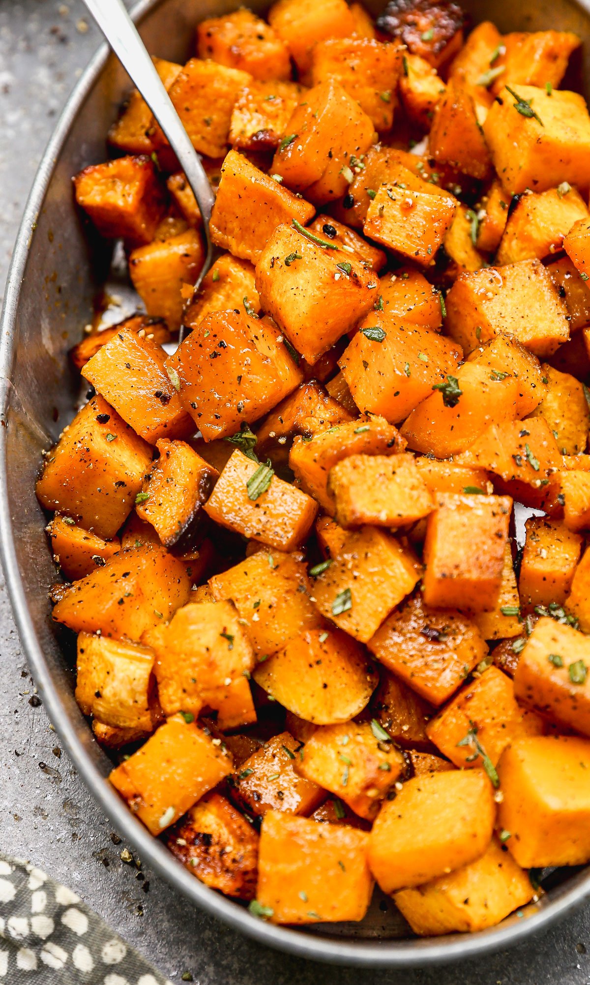 A close-up of roasted butternut squash cubes seasoned with herbs and pepper in an oval dish with a spoon.