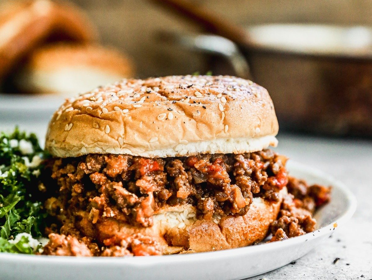 A sloppy joe sandwich with seasoned ground beef on a sesame bun, served on a white plate with a side of greens.
