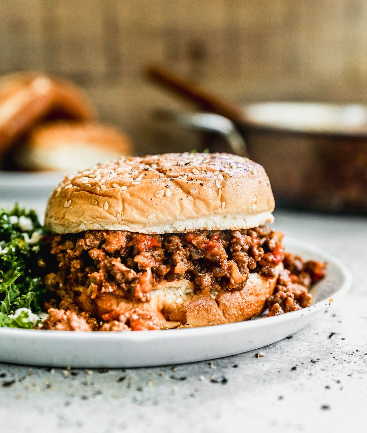 A sloppy joe sandwich with seasoned ground beef on a sesame bun, served on a white plate with a side of greens.