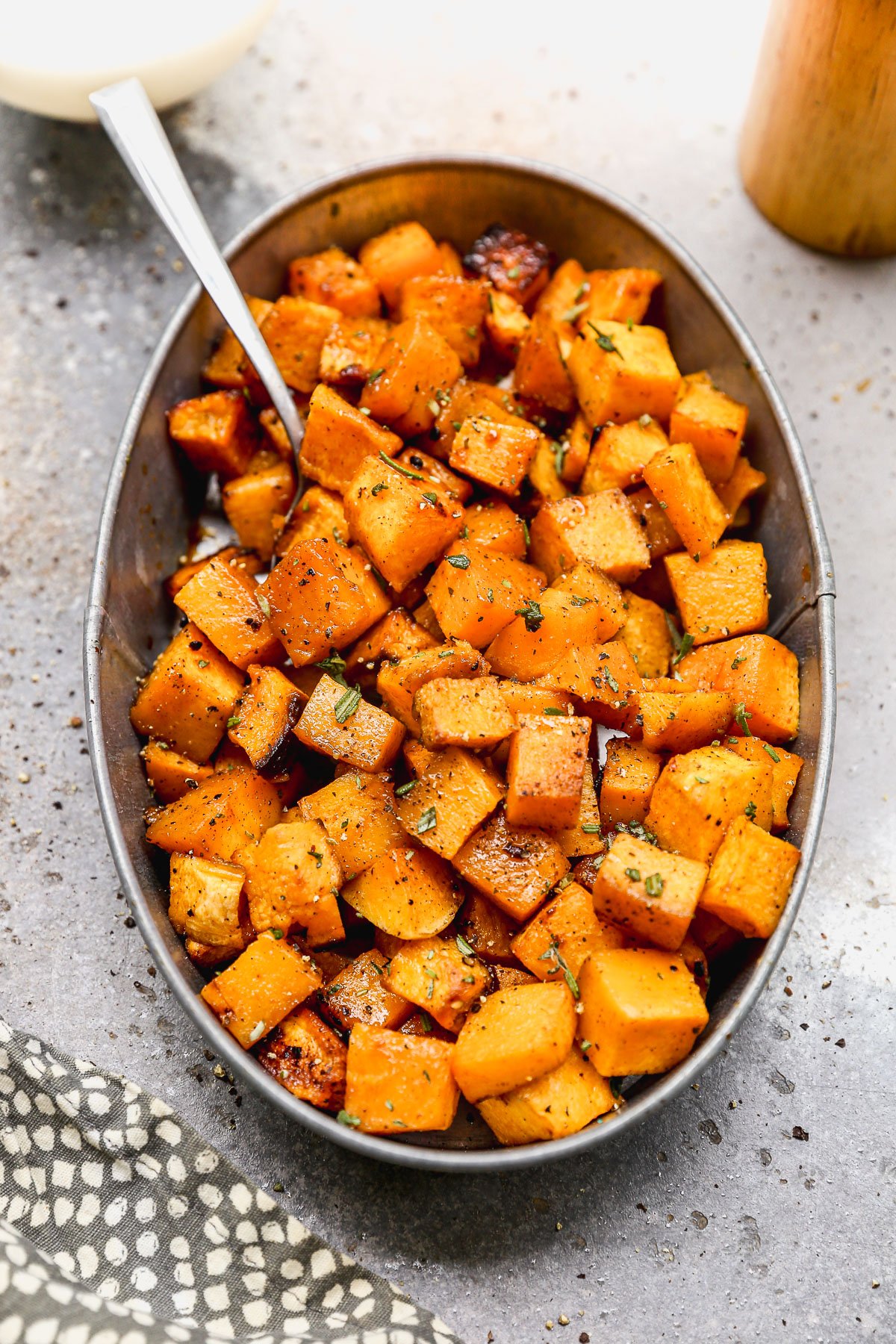 Oval dish filled with roasted, seasoned butternut squash cubes, with a spoon, on a gray countertop.