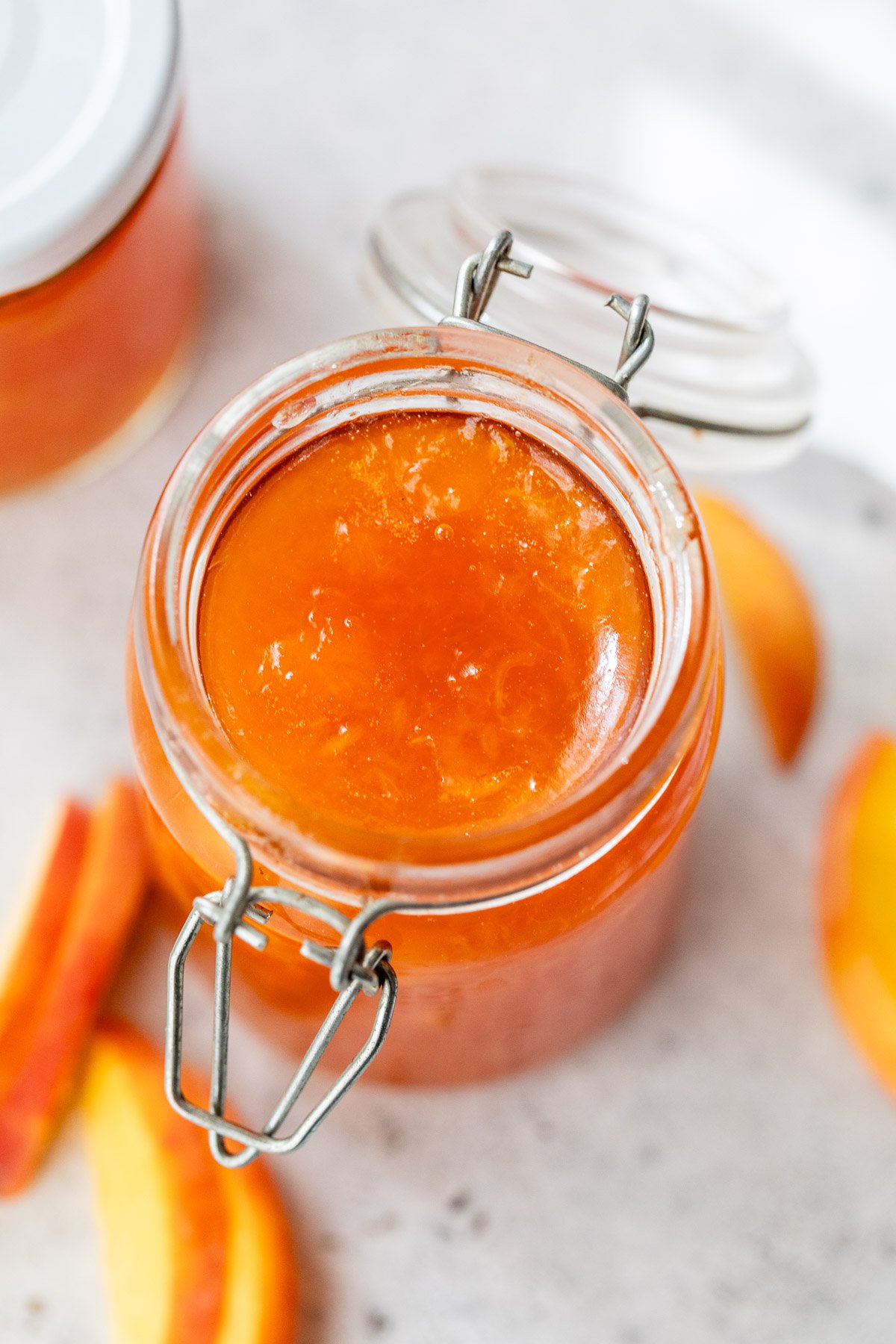 Open glass jar filled with peach jam, surrounded by peach slices on a light surface.