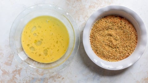 A glass bowl of beaten eggs next to a white dish of seasoned breadcrumbs on a light surface.