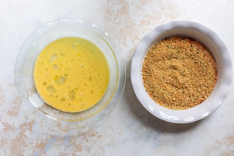 A glass bowl of beaten eggs next to a white dish of seasoned breadcrumbs on a light surface.
