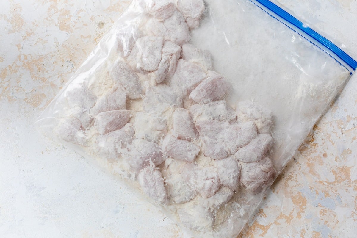 Raw, flour-coated chicken pieces in a clear plastic zip-top bag on a light textured surface.