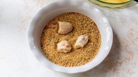 Three pieces of raw chicken coated in breadcrumbs in a white bowl on a light surface.