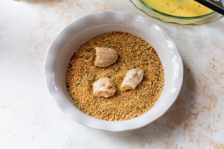 Three pieces of raw chicken coated in breadcrumbs in a white bowl on a light surface.