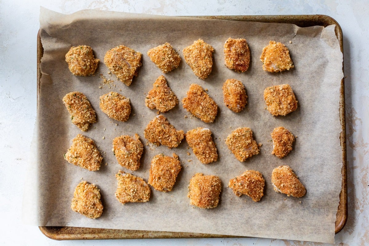 A baking tray lined with parchment paper holds evenly spaced, breaded chicken nuggets.
