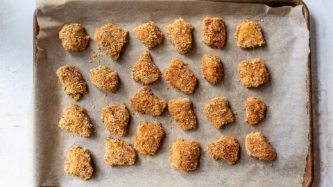 A baking tray lined with parchment paper holds evenly spaced, breaded chicken nuggets.