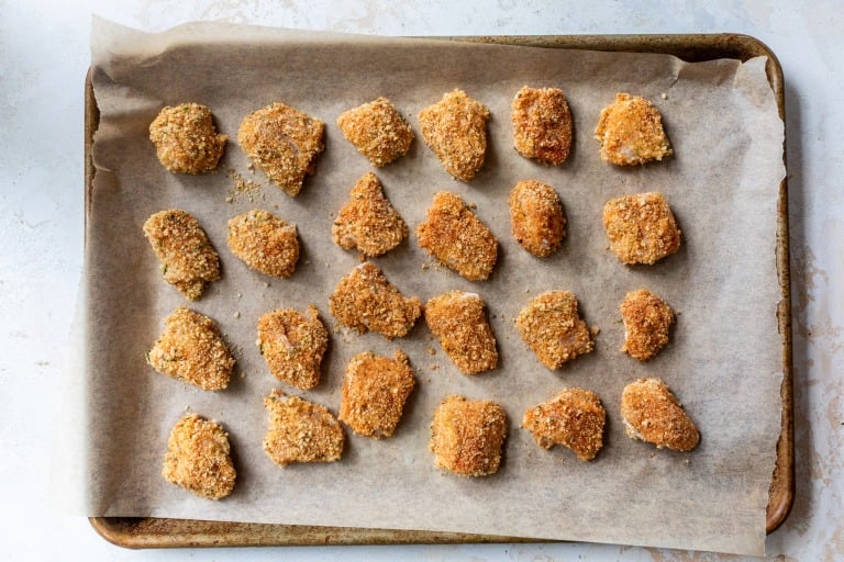 A baking tray lined with parchment paper holds evenly spaced, breaded chicken nuggets.