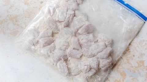 Raw, flour-coated chicken pieces in a clear plastic zip-top bag on a light textured surface.