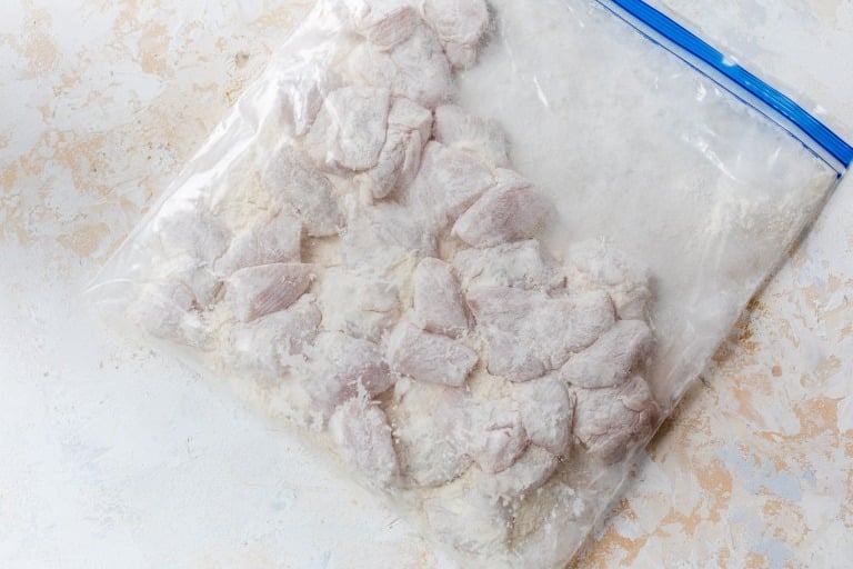 Raw, flour-coated chicken pieces in a clear plastic zip-top bag on a light textured surface.