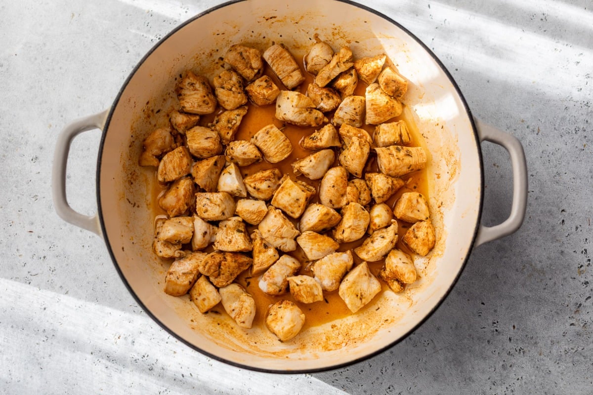 Cubed, seasoned chicken pieces cooking in a round, white pot on a light gray countertop.
