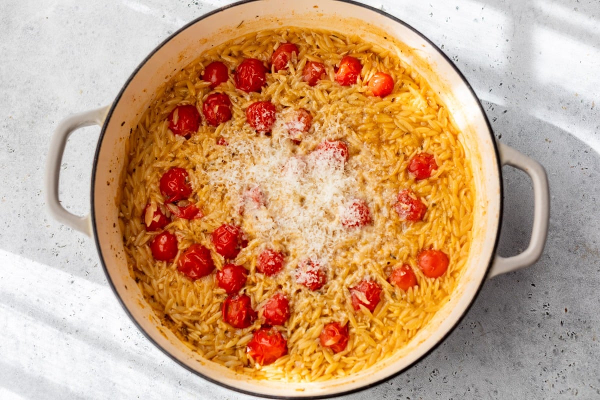 A pot of cooked orzo pasta with cherry tomatoes and grated cheese on top, sitting on a light-colored surface.