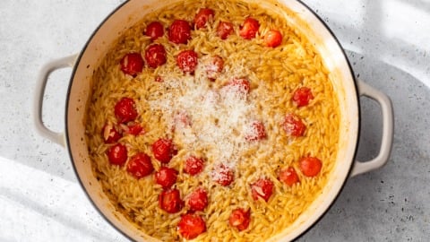 A pot of cooked orzo pasta with cherry tomatoes and grated cheese on top, sitting on a light-colored surface.