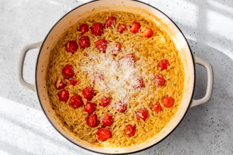 A pot of cooked orzo pasta with cherry tomatoes and grated cheese on top, sitting on a light-colored surface.
