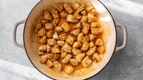 Cubed, seasoned chicken pieces cooking in a round, white pot on a light gray countertop.