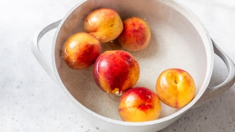 Six nectarines in a pot of water on a light gray surface.