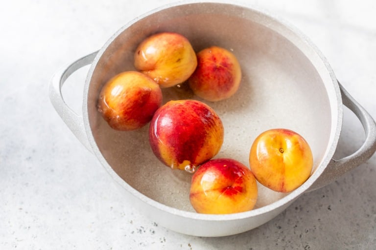 Six nectarines in a pot of water on a light gray surface.
