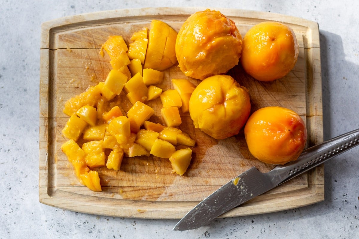 Peeled peaches and diced peach pieces on a wooden cutting board, with a knife beside them.
