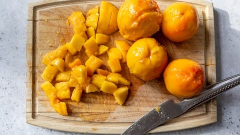 Peeled peaches and diced peach pieces on a wooden cutting board, with a knife beside them.