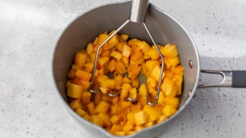A potato masher mashes chopped peaches in a gray saucepan on a light-colored countertop.