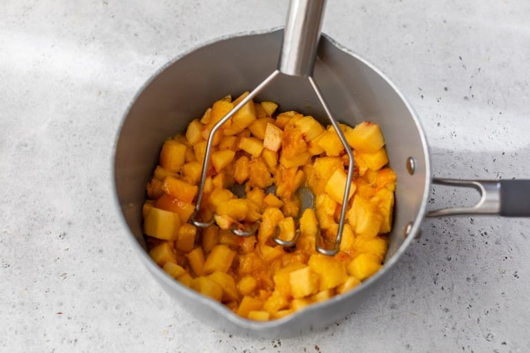 A potato masher mashes chopped peaches in a gray saucepan on a light-colored countertop.