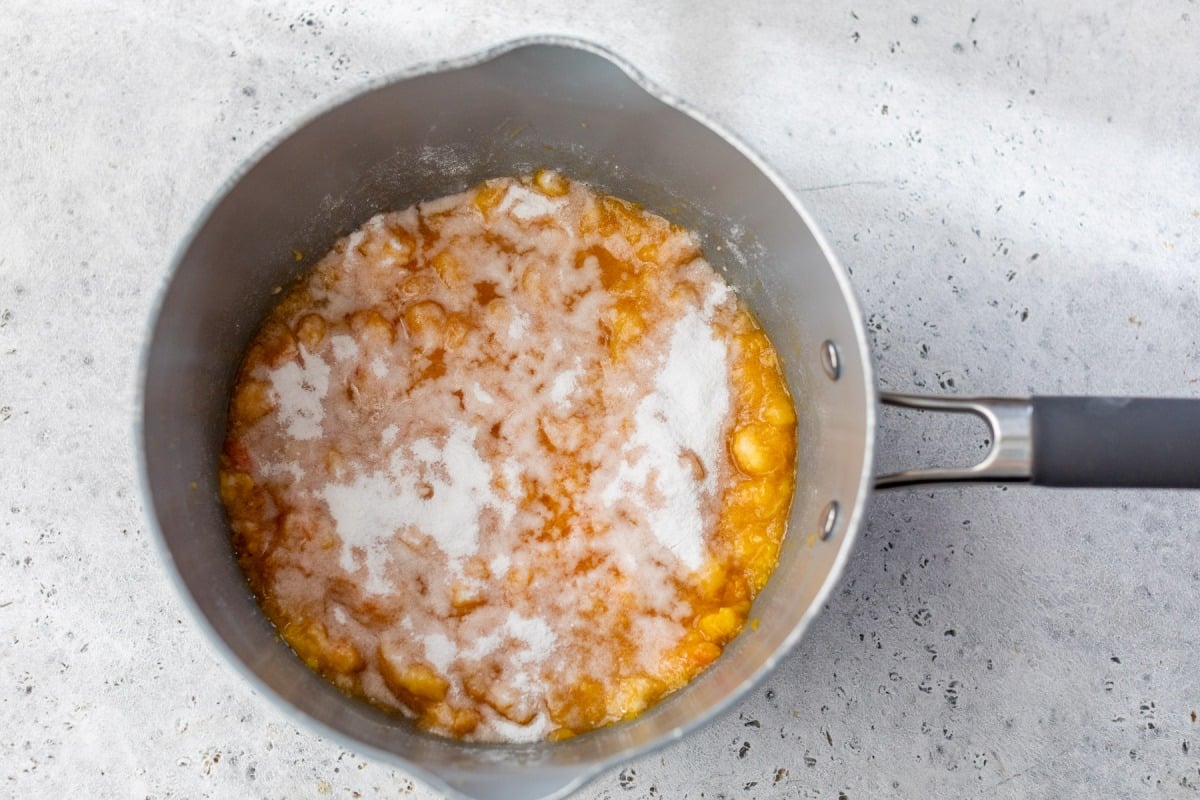 A saucepan filled with peaches and a layer of white sugar on top, on a light countertop.