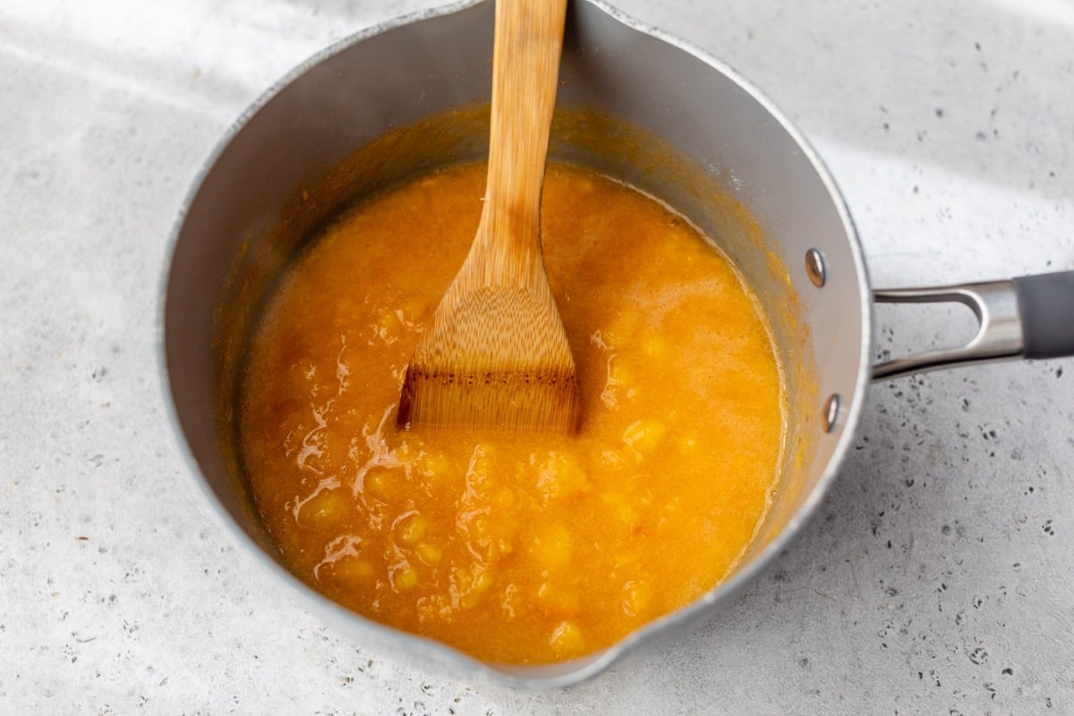 A saucepan with peach jam and a wooden spatula stirring it on a light gray countertop.