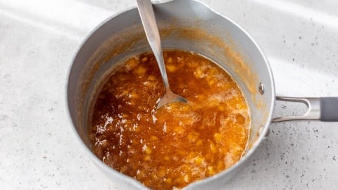 A saucepan with peach jam being stirred with a metal spoon on a light countertop.