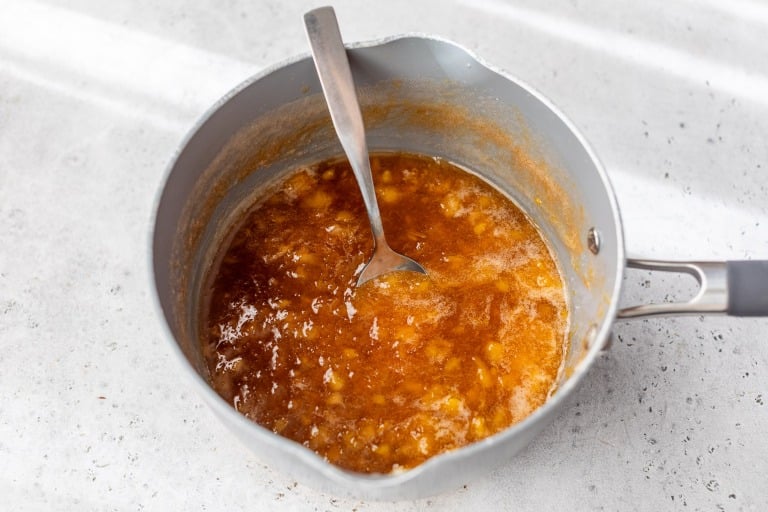 A saucepan with peach jam being stirred with a metal spoon on a light countertop.