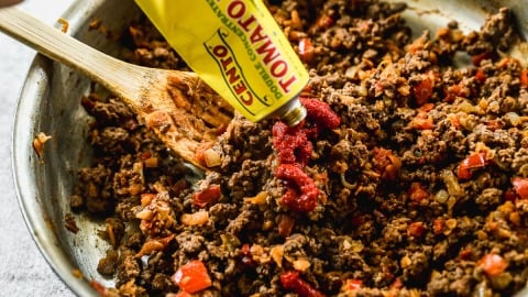 A hand squeezes tomato paste from a tube into a skillet of cooked ground beef and vegetables being stirred with a wooden spoon.